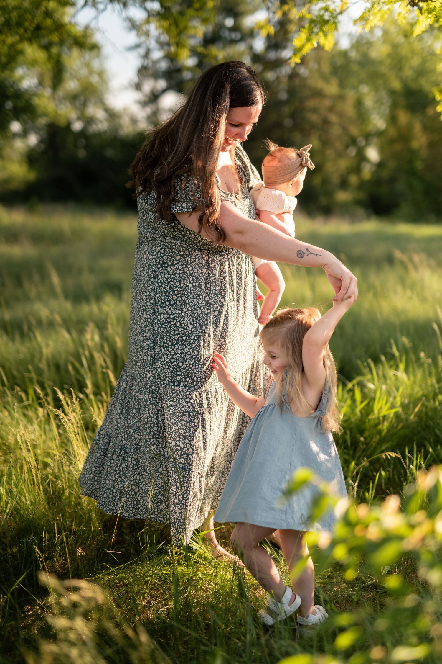 Mother in floral dress holds baby while young daughter dances in sunlit green meadow.