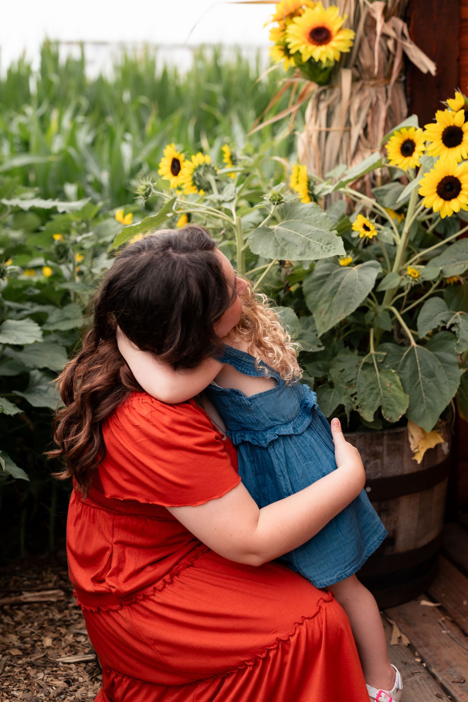 Mother in red dress hugging young child in denim dress among vibrant sunflowers in a garden setting.