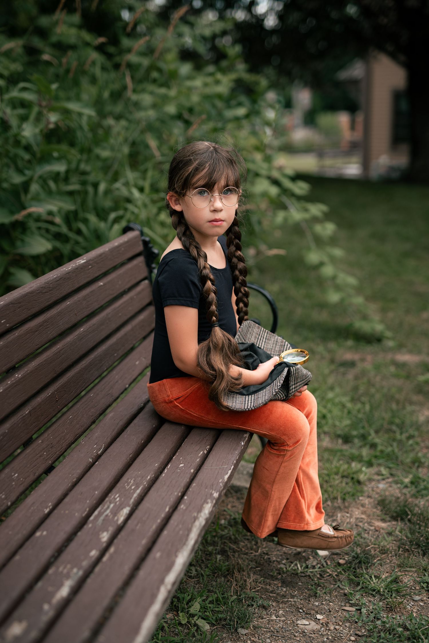 Young girl with braided hair and glasses sits on a wooden park bench wearing orange pants and black top.