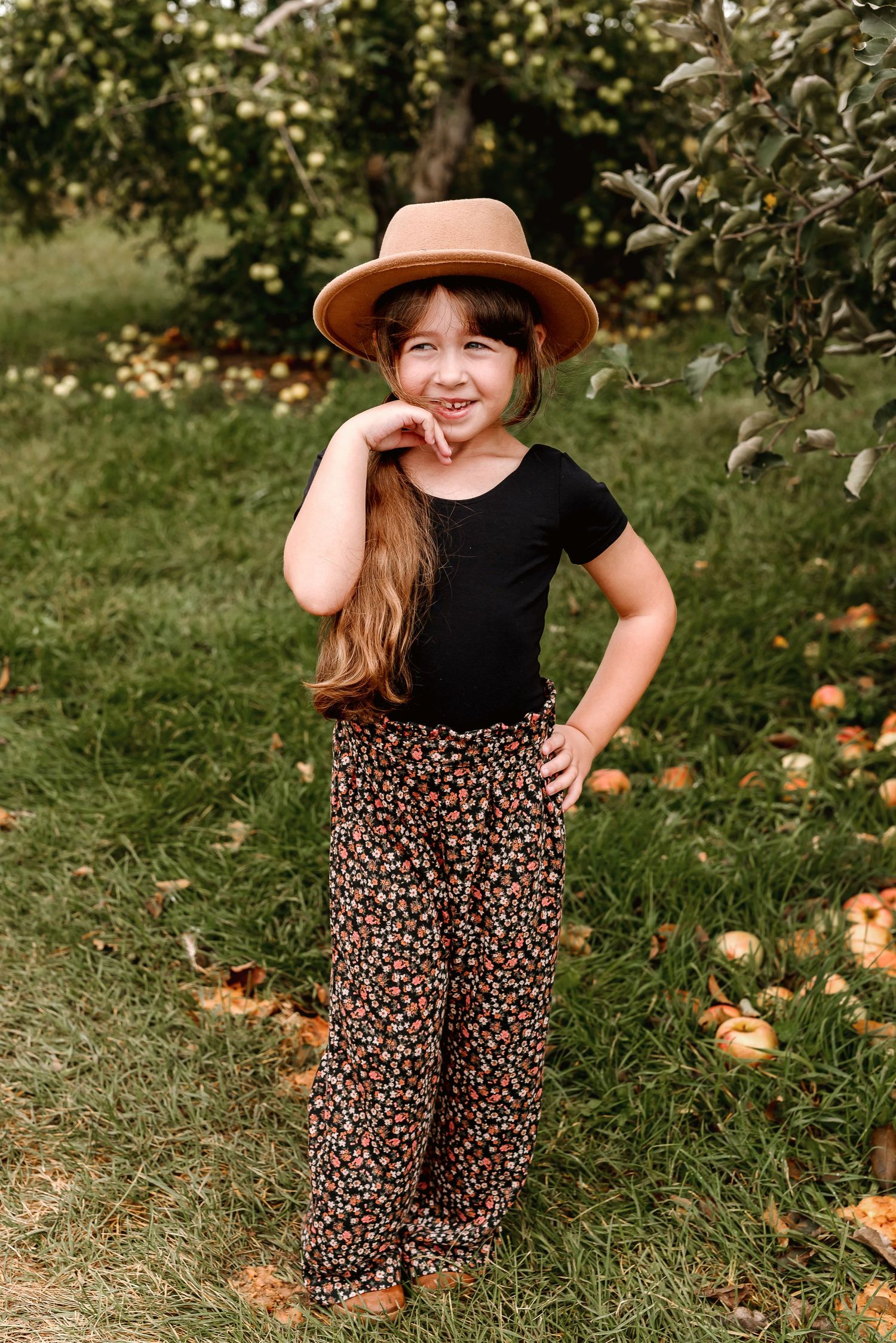A person wearing a black top and floral skirt poses playfully in a garden with fallen pumpkins on the grass.