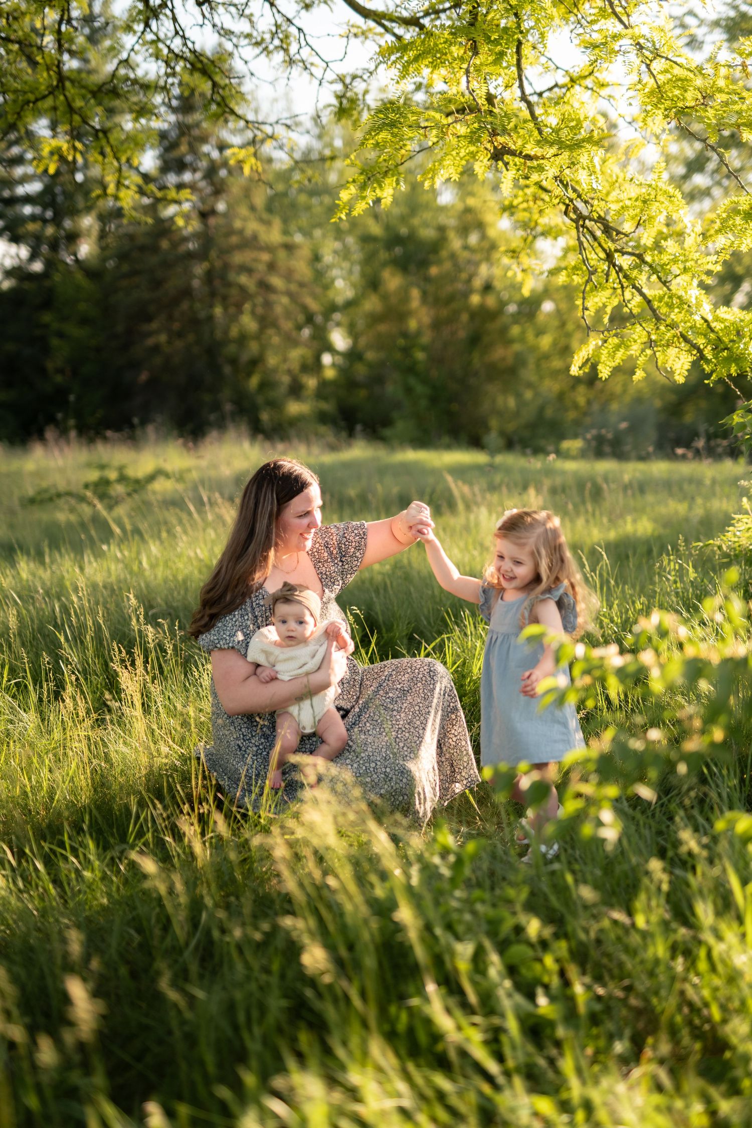 Mother holding baby while dancing with toddler daughter in sunny green meadow during golden hour.