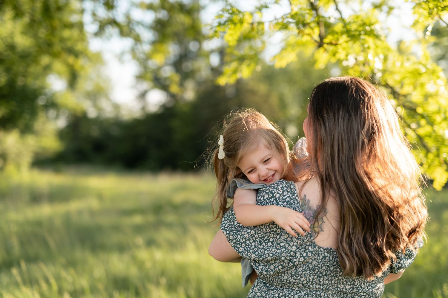 A mother holds and hugs her laughing little girl outdoors in a sunny green park, both wearing floral dresses.