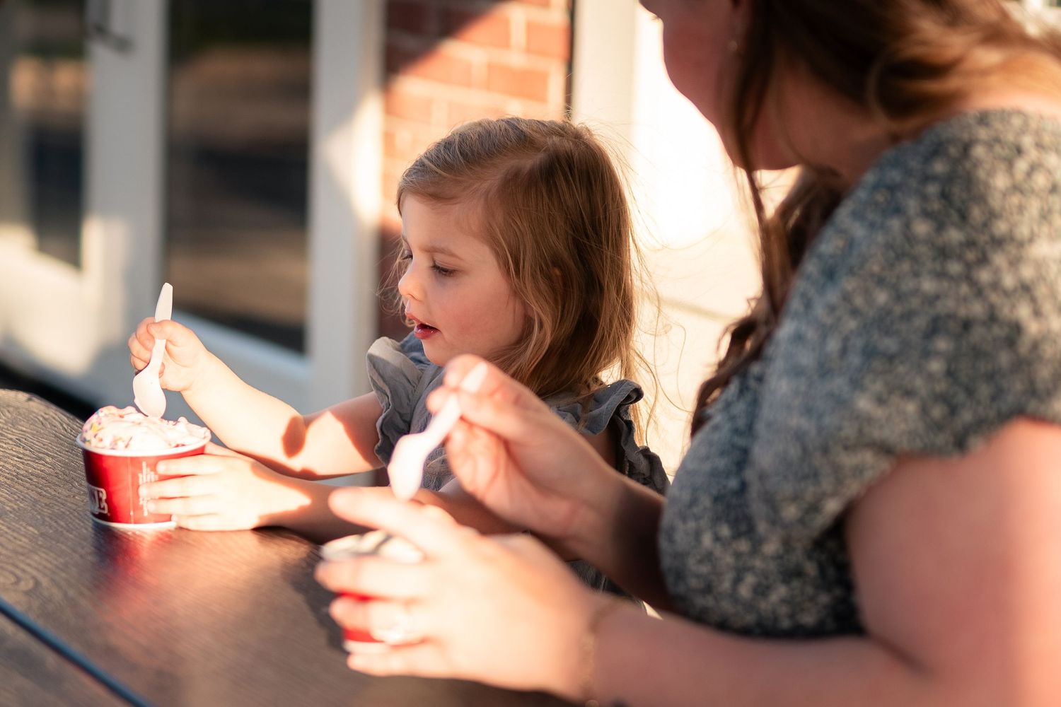 A young girl and woman enjoy ice cream together outdoors in warm golden sunlight.