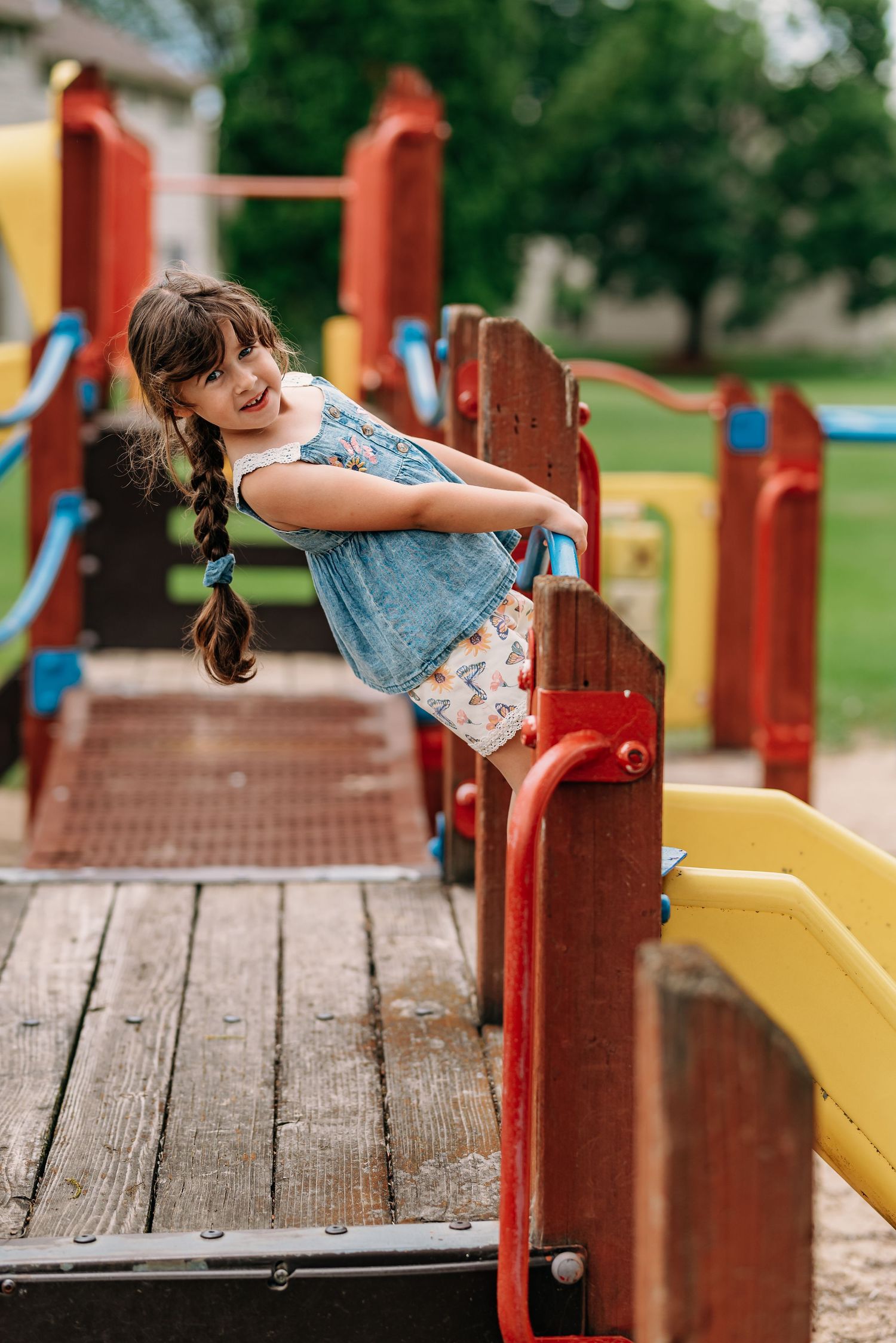 A child plays on colorful wooden playground equipment on a summer day, leaning over a red railing.