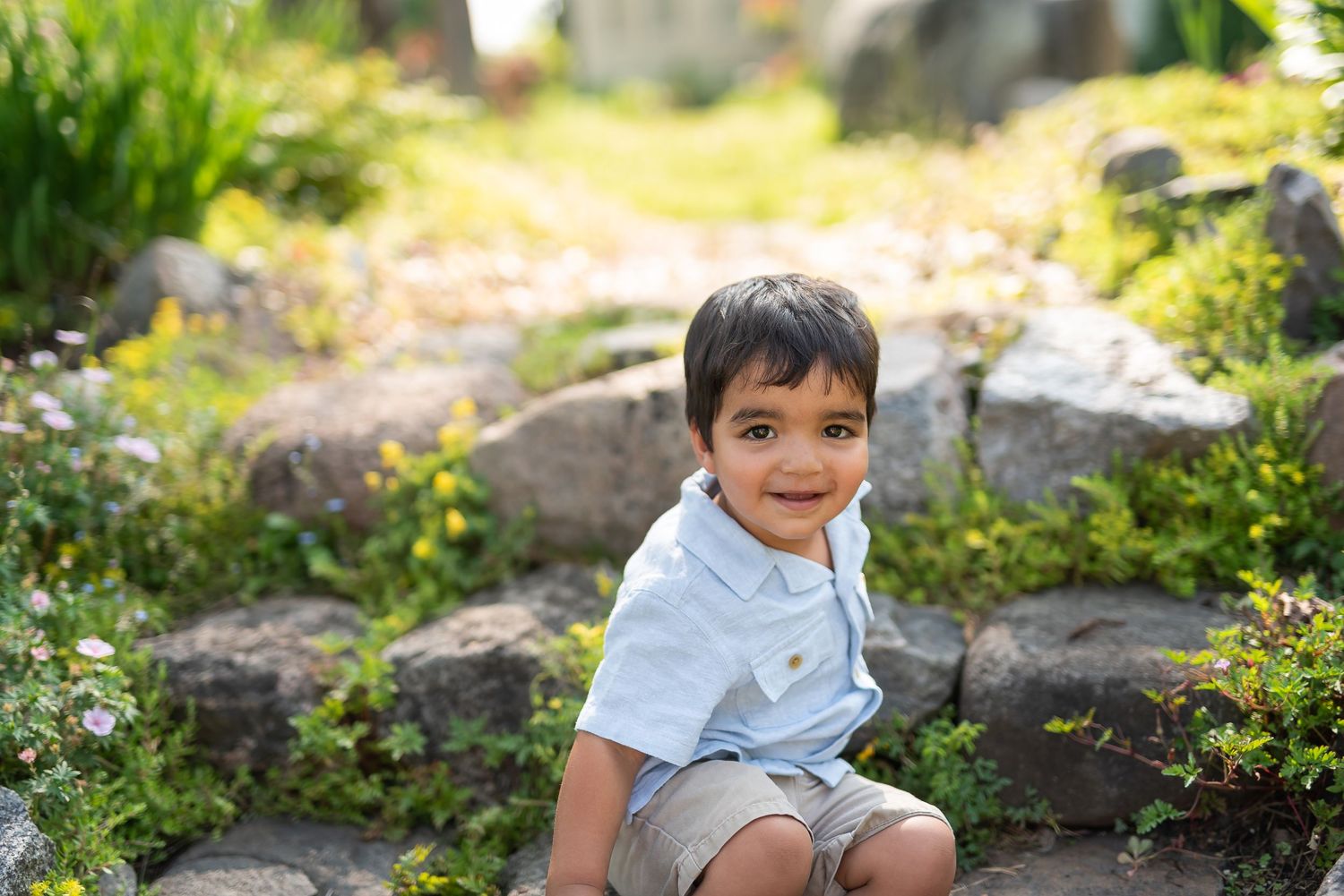 A smiling young boy in a light blue shirt and khaki shorts crouching in a lush green garden with rocks.