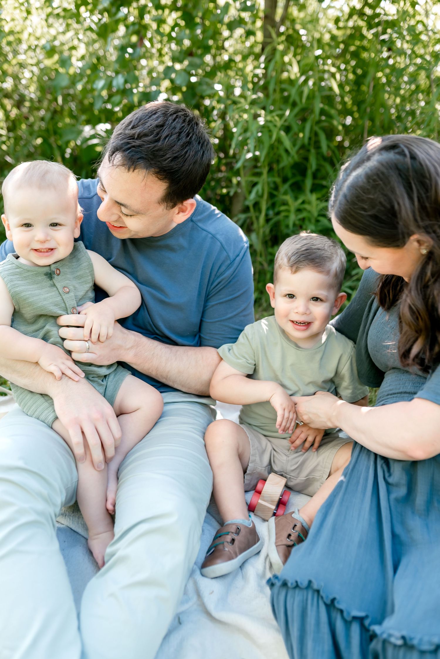 Happy family of four sitting outdoors, parents smiling at two young boys in coordinating blue and green outfits.