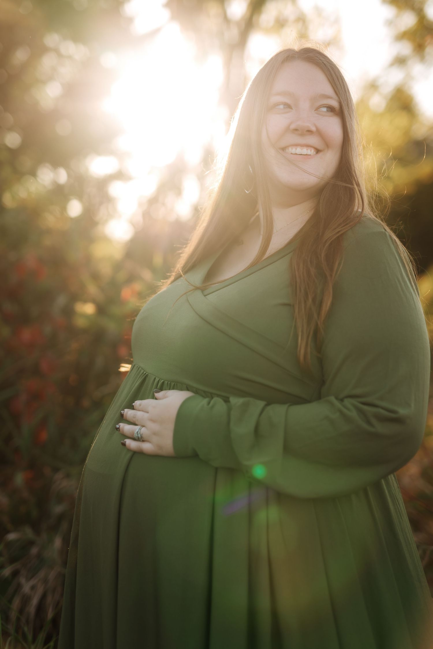 A pregnant woman in a green dress stands in golden sunlight during a maternity photoshoot outdoors.
