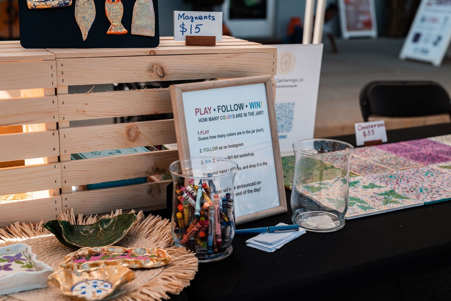Outdoor market vendor table displaying magnets for $15, crayons, and a play and follow sign with wooden crates.