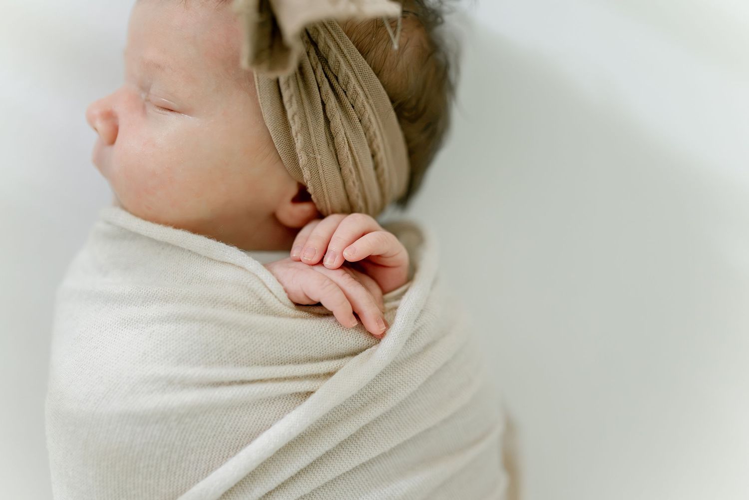 A peacefully sleeping newborn baby swaddled in a cream blanket with a tan bow headband.