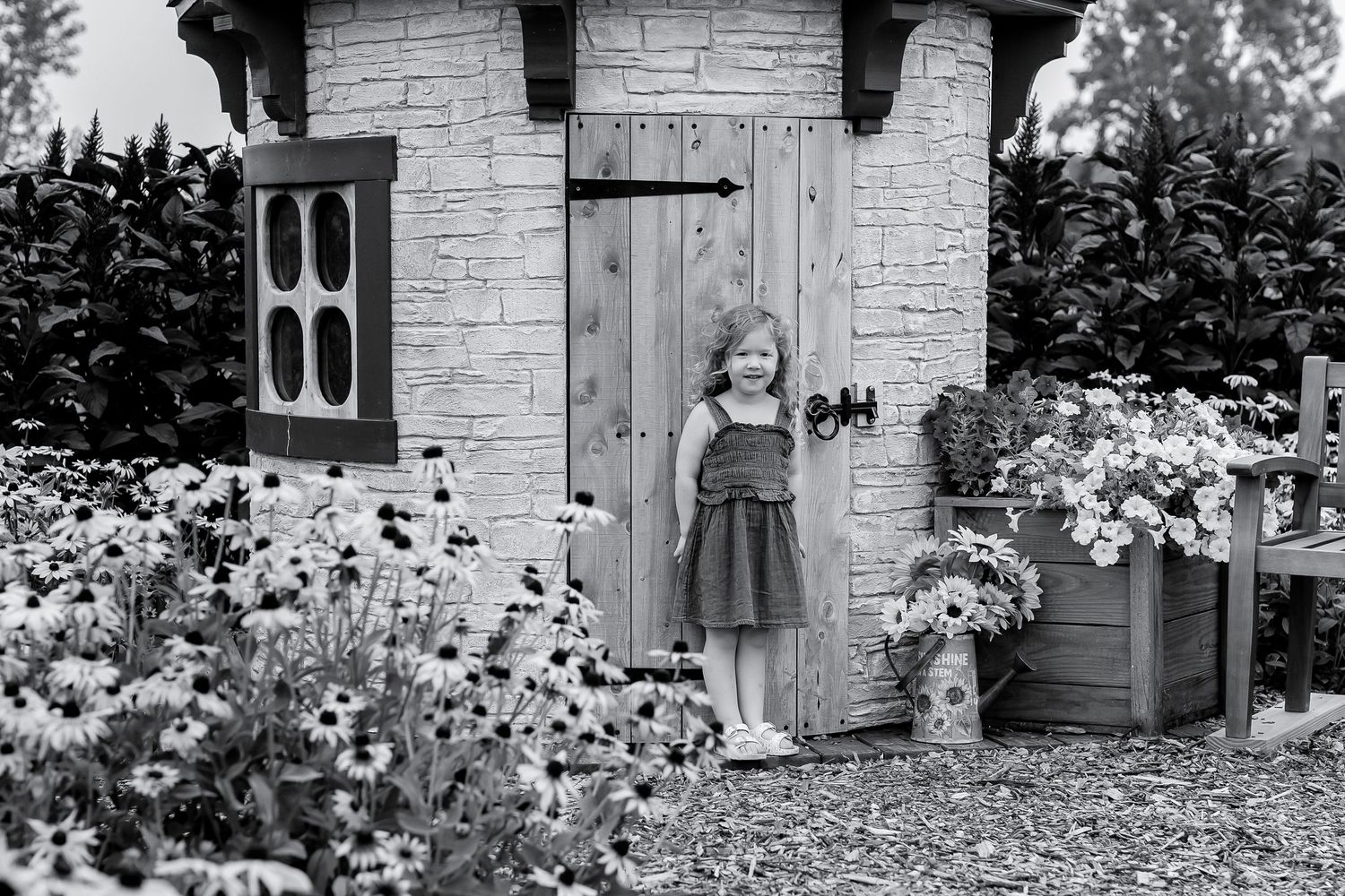 Young girl standing by a charming stone playhouse surrounded by blooming flowers in a garden.