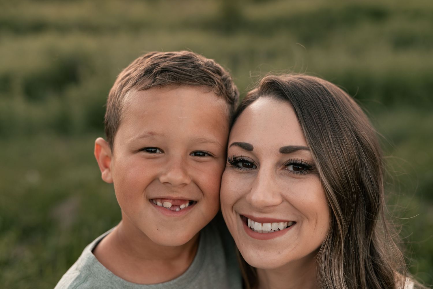 A smiling boy and young woman pose cheek-to-cheek outdoors in a green field for a family portrait.