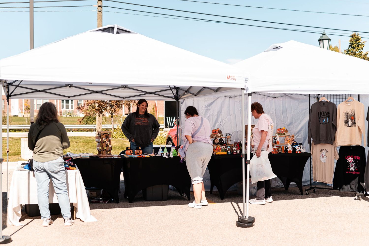 Vendors and shoppers browse colorful merchandise under white canopy tents at an outdoor art festival.