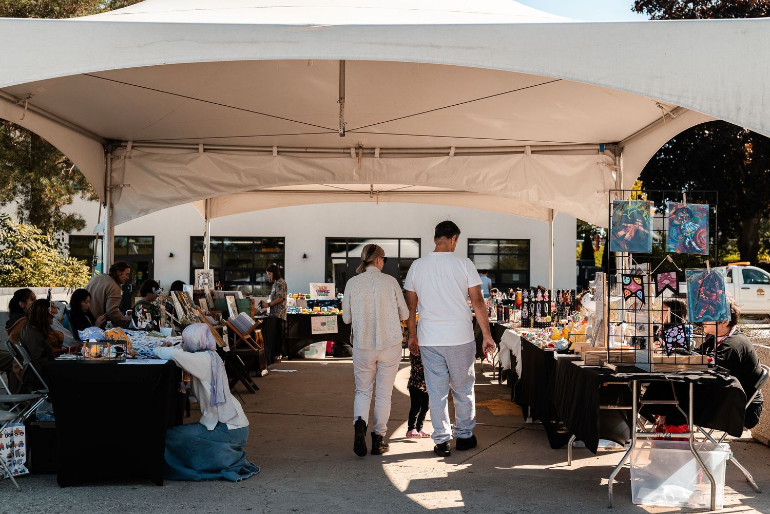 Outdoor market under white tent with vendors and shoppers browsing art and goods in sunlight.