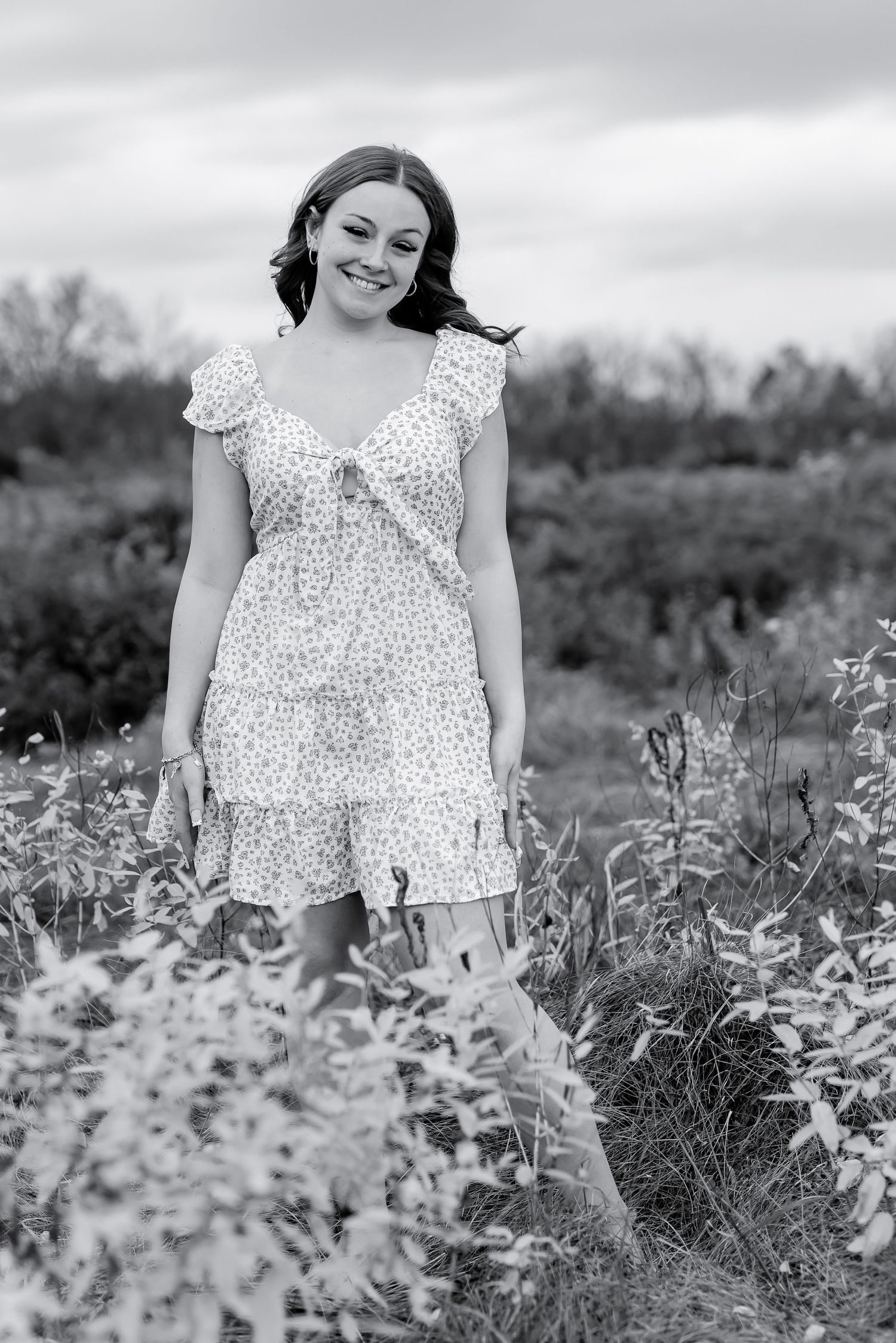 Black and white photo of smiling young woman in floral dress standing in autumn wildflower field.