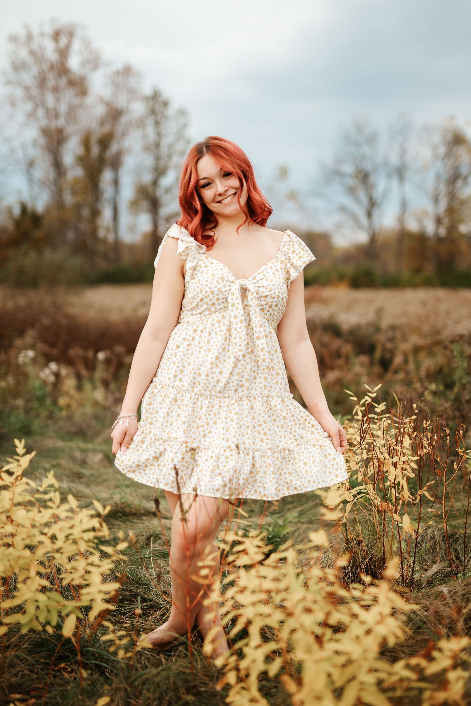 Smiling redhead woman in floral sundress standing in autumn field with golden wildflowers.