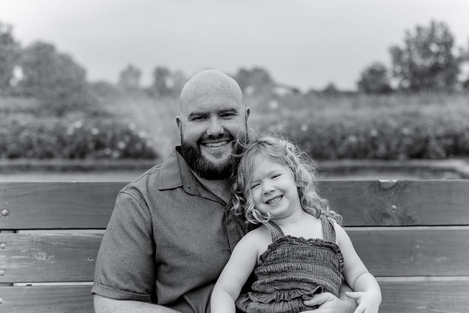 Black and white photo of smiling bald father and curly-haired daughter on bench.
