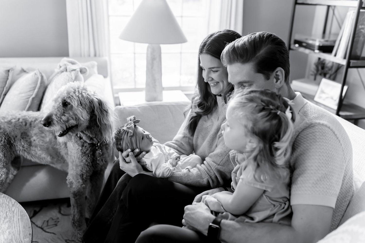 Black and white photo of family with newborn, toddler, and fluffy dog on living room sofa.