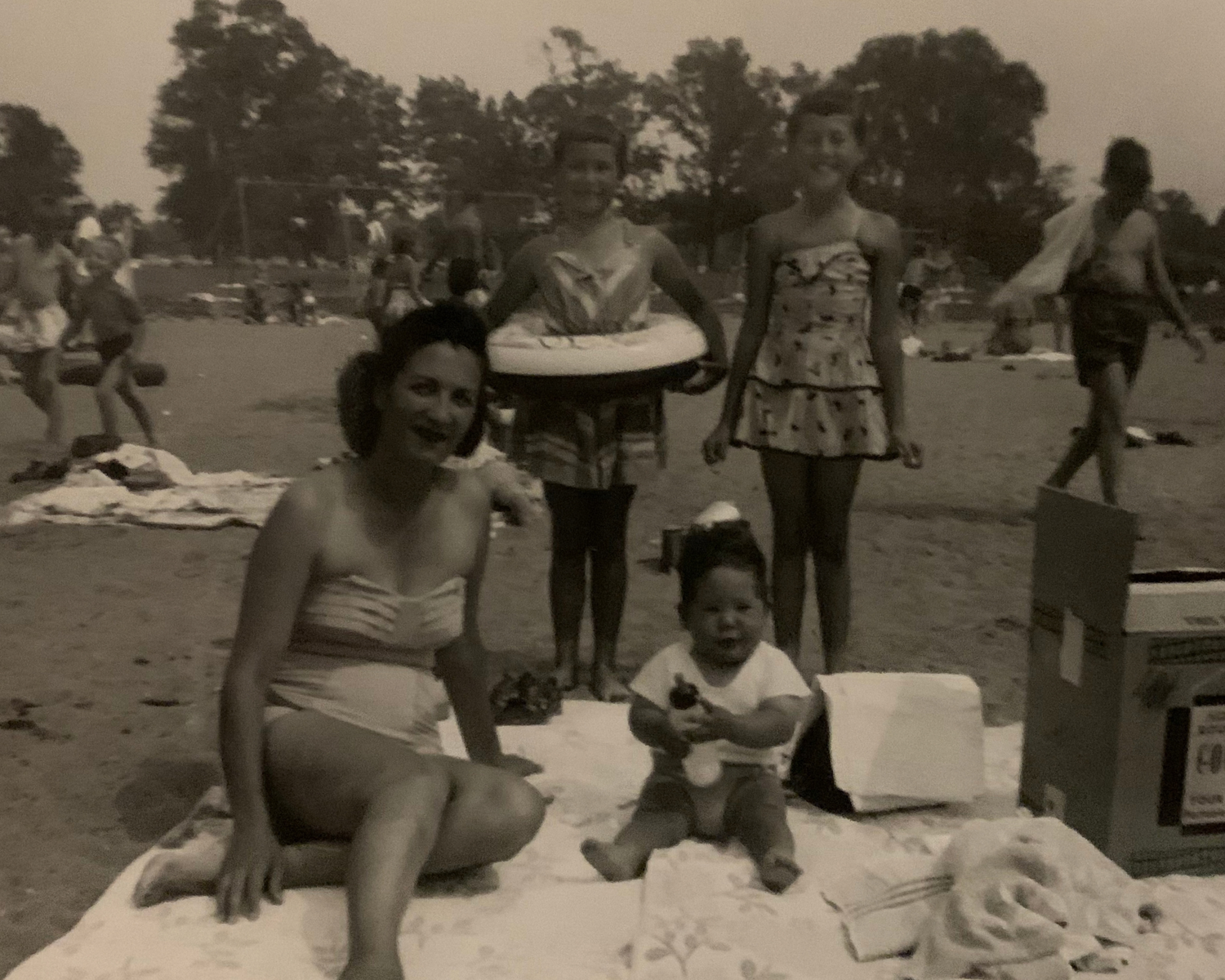 Vintage black and white beach photo of a family with two women, a child, and a baby sitting on a blanket in the sand.