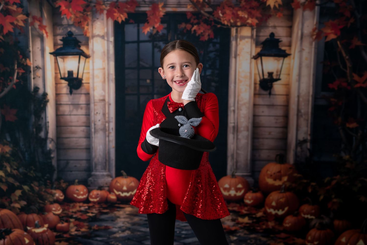Girl in red dress holds magic hat with rabbit on Halloween backdrop with pumpkins and autumn leaves.