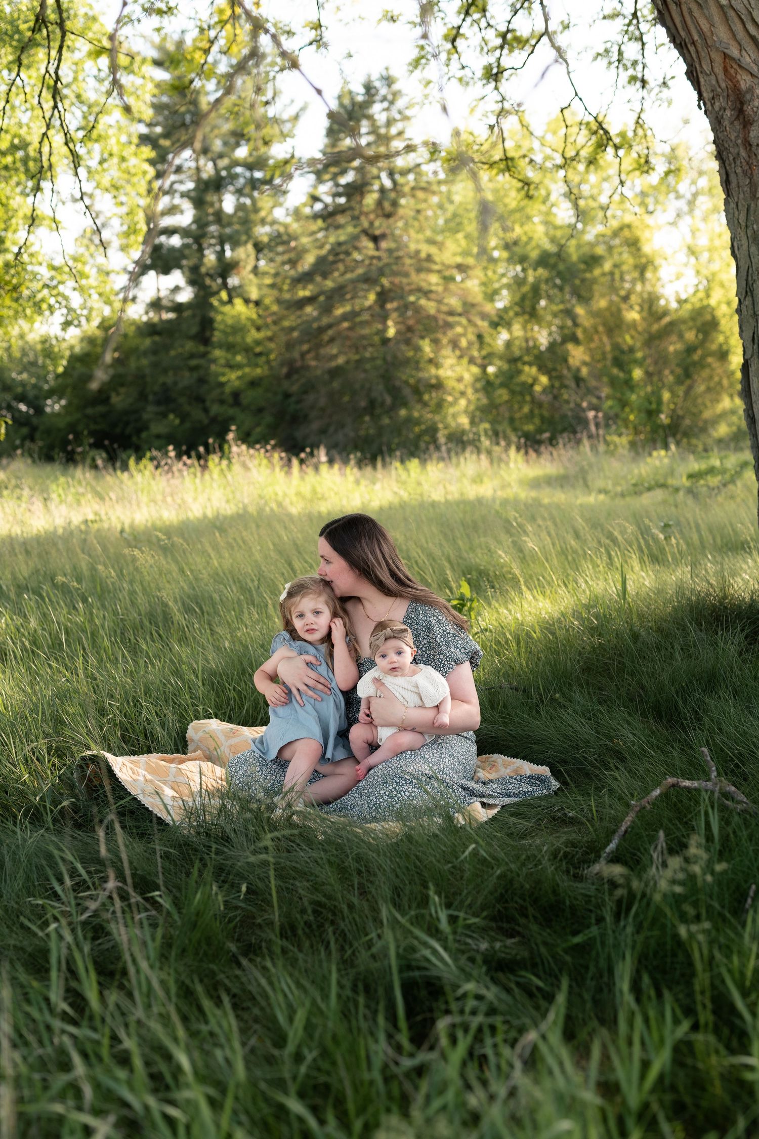 Mother kissing toddler while holding baby on blanket in sunny green meadow with trees.