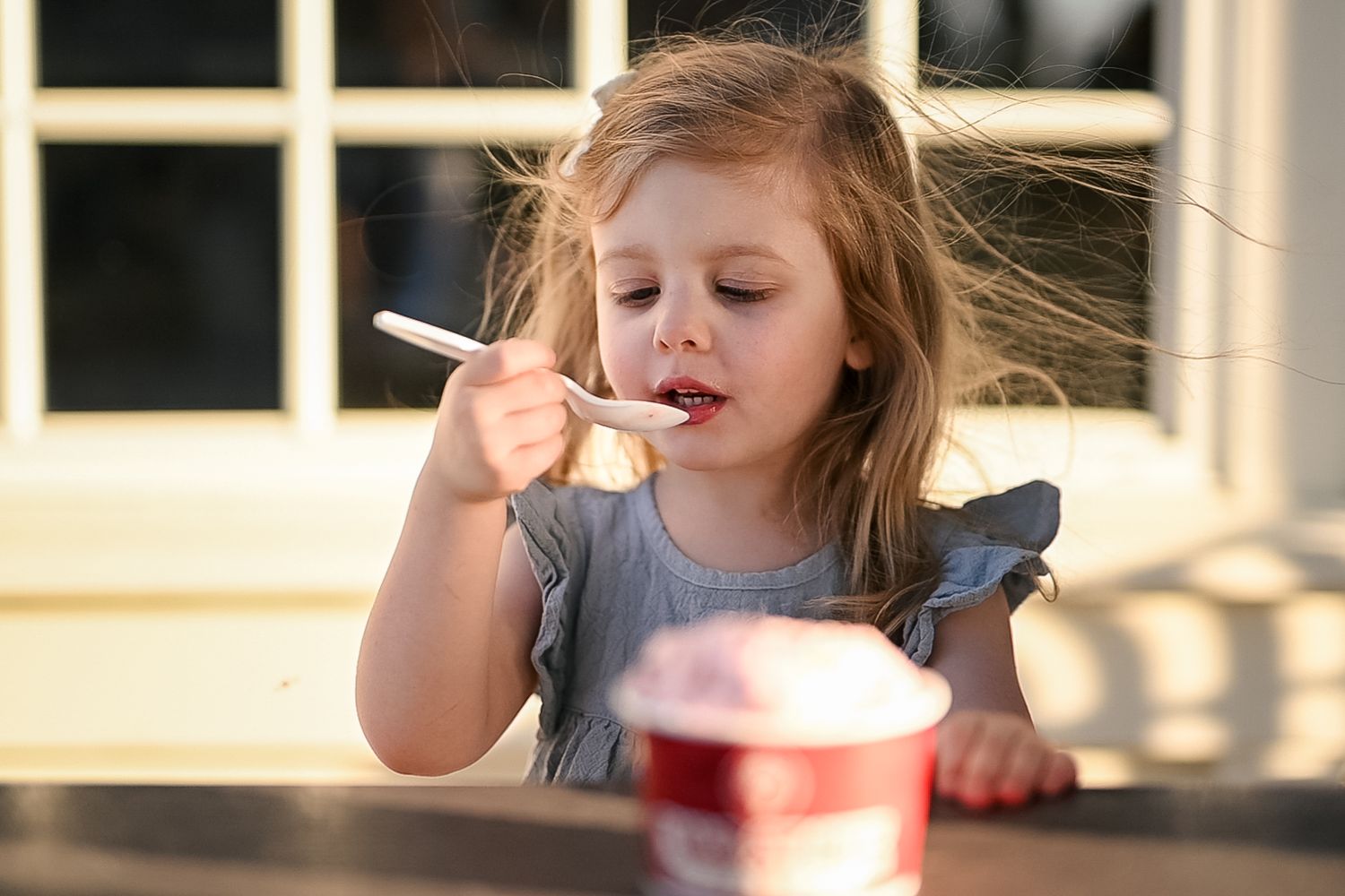 Young girl eating yogurt with a spoon from a red cup outdoors in warm sunlight.