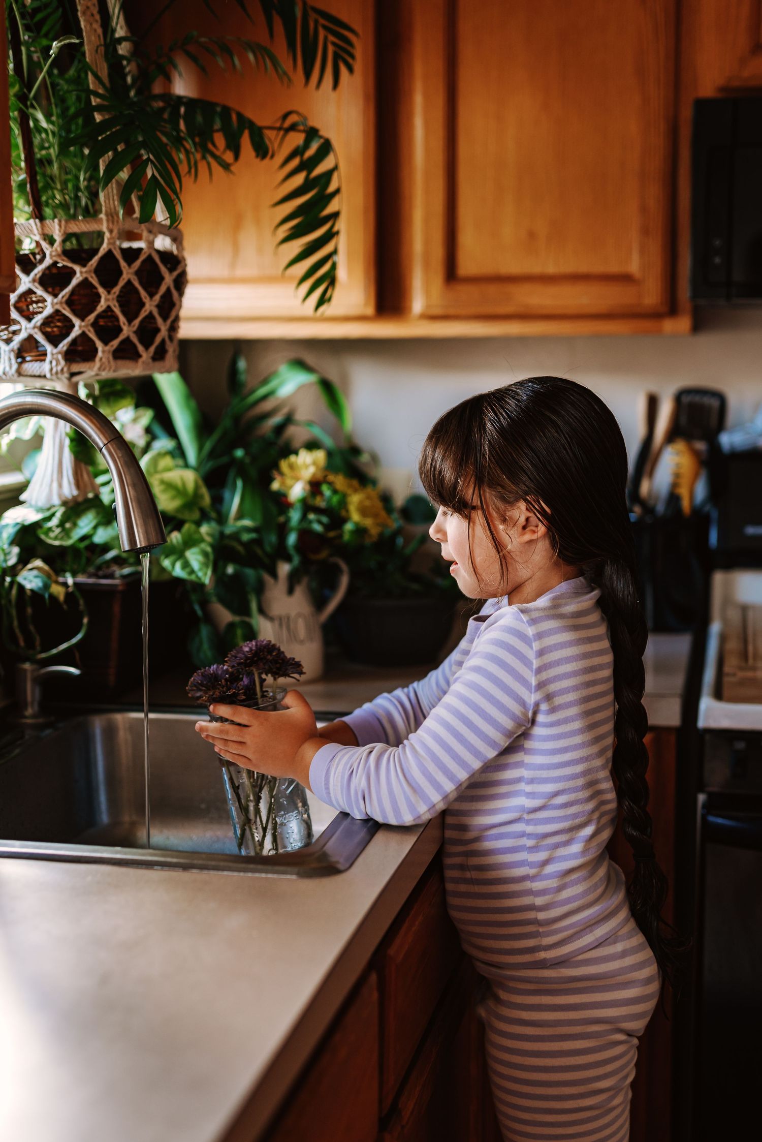 Child in striped shirt stands at kitchen counter examining houseplants in morning light.