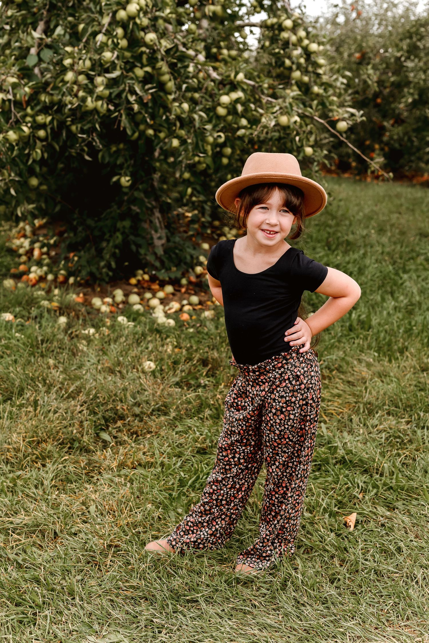 A person in a brown hat and floral dress sits in a grassy orchard with fallen fruit scattered on the ground.