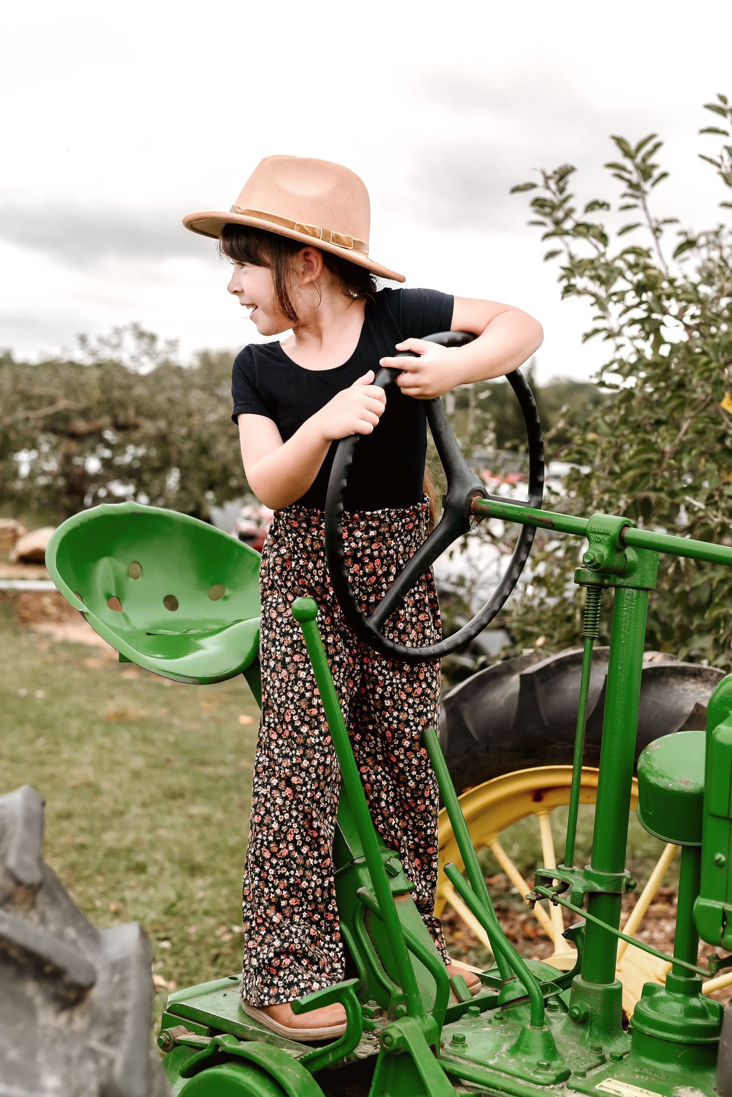 Someone wearing a leopard print dress and sun hat sits on a vintage green tractor in a rural farm setting.
