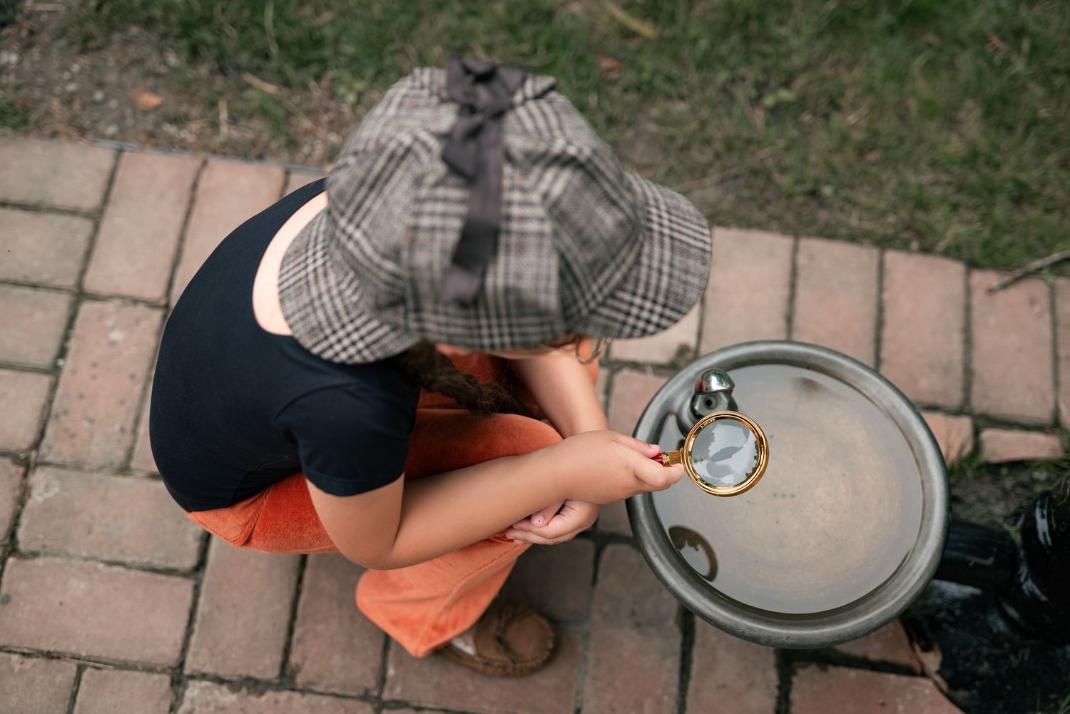 Child wearing plaid detective hat crouches on brick path, examining a magnifying glass over a silver tray.