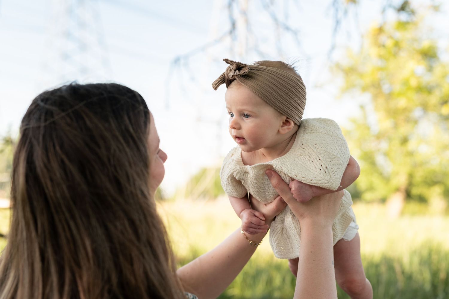 A mother lifts her adorable baby girl wearing a bow headband outdoors in a sunny green park setting.