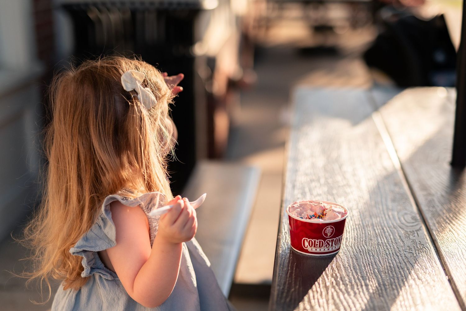 Young girl with long hair sitting at a wooden table with a red Rita's Italian Ice cup outdoors.