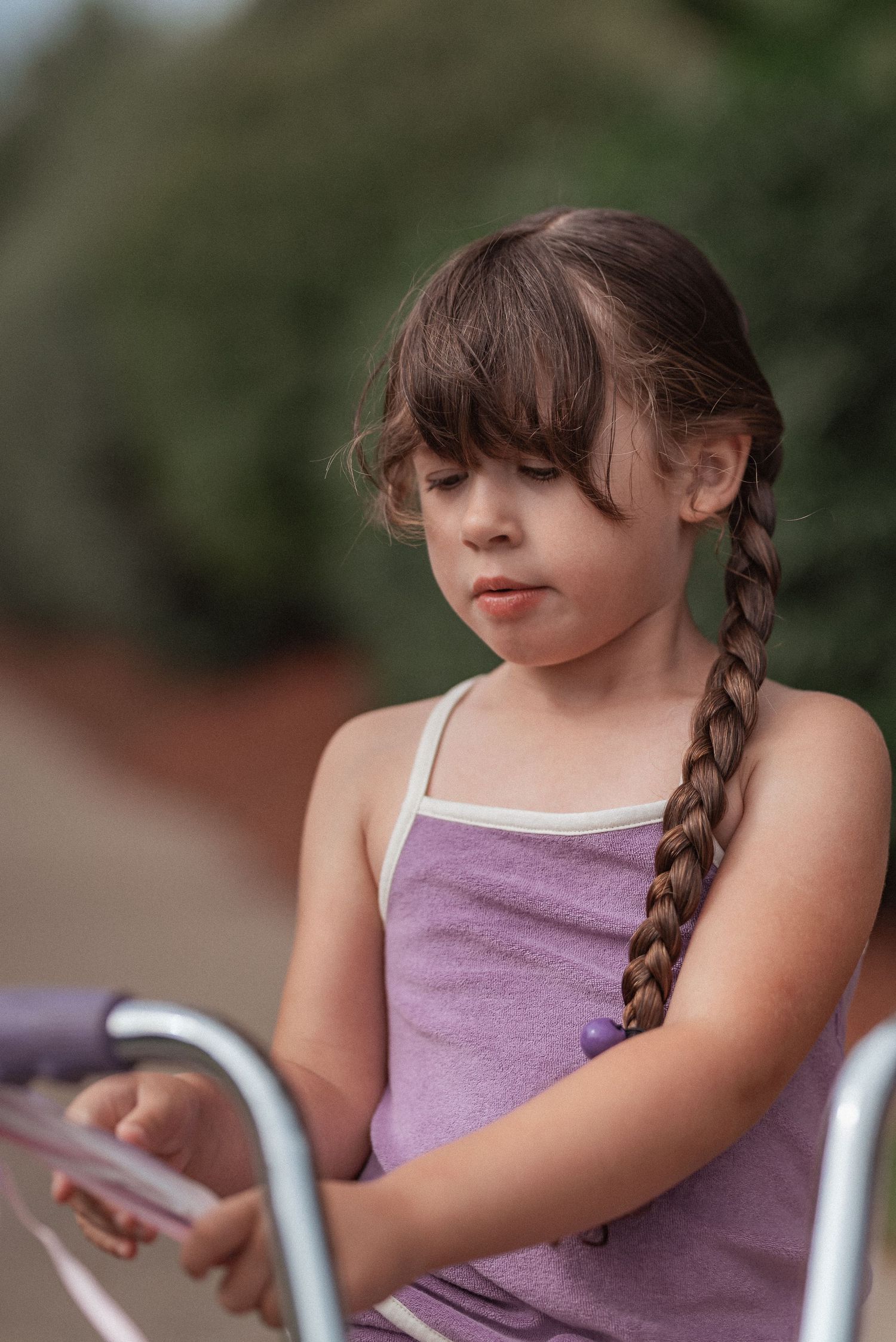 A braided-hair child in a purple tank top sitting on a bicycle and looking downward.