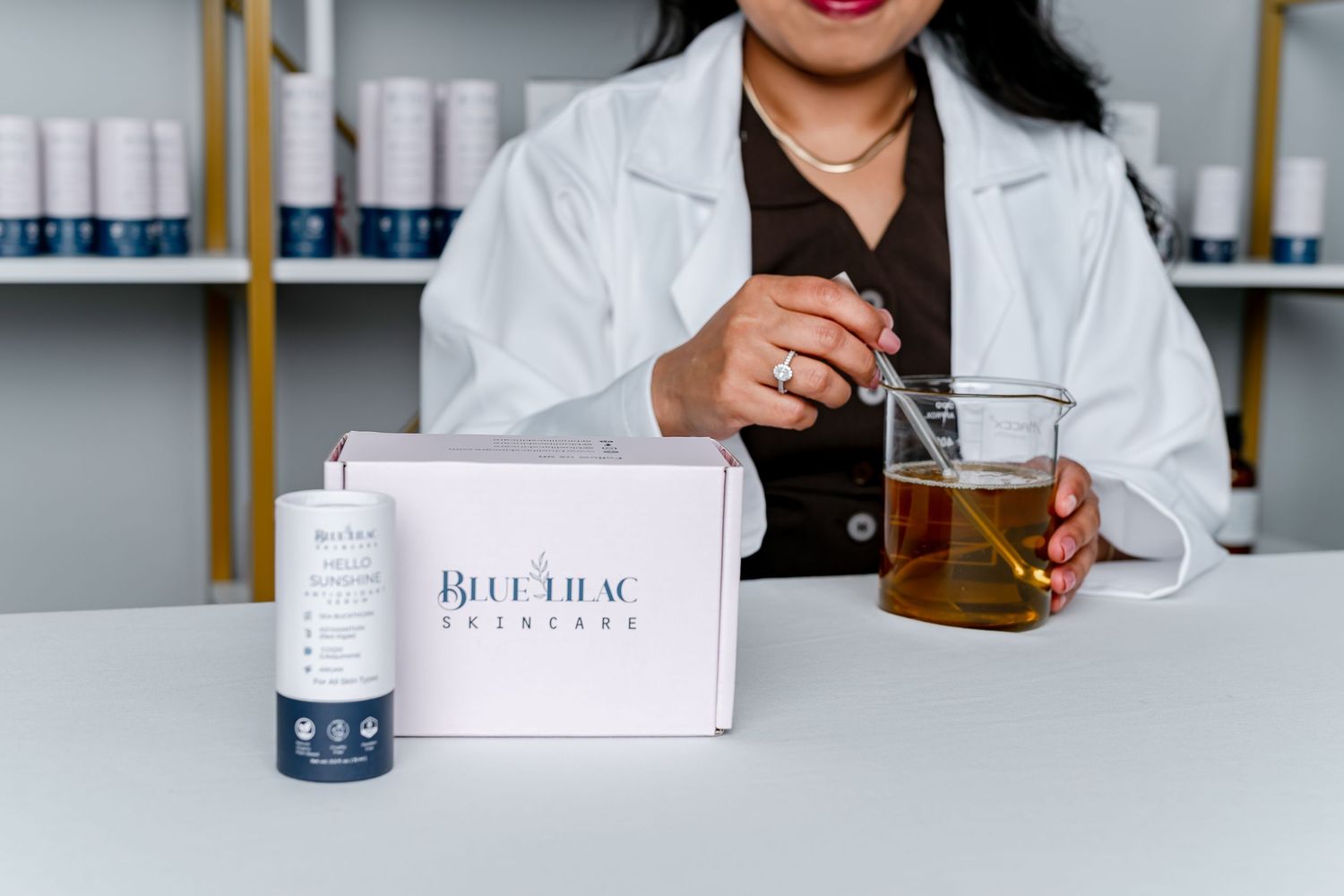 A pharmacist in a white lab coat holds tea beside a Blue Lilac Skincare box on a white table.