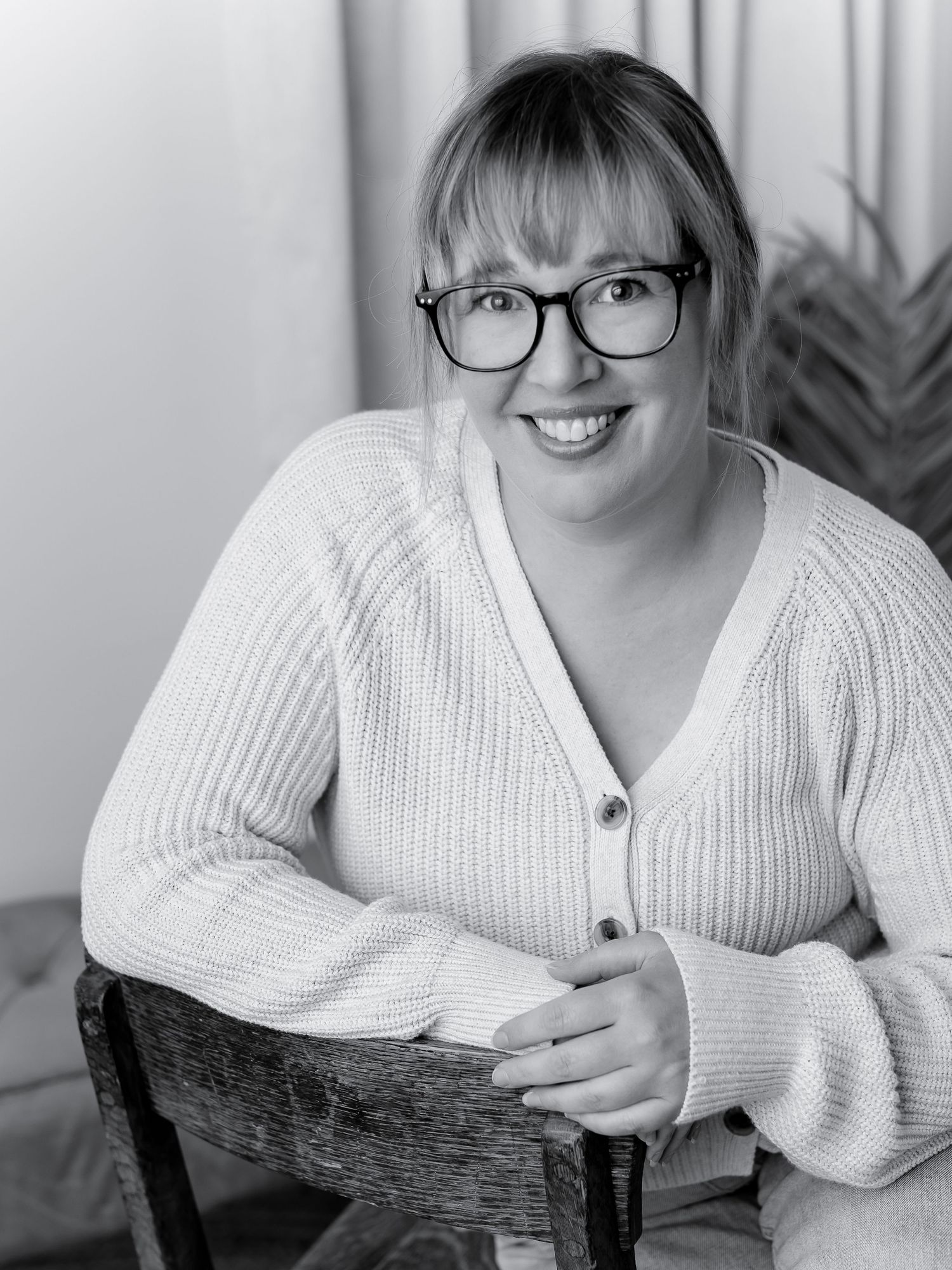 Black and white portrait of a smiling woman with bangs and glasses, wearing a knit cardigan, seated on a wooden chair.