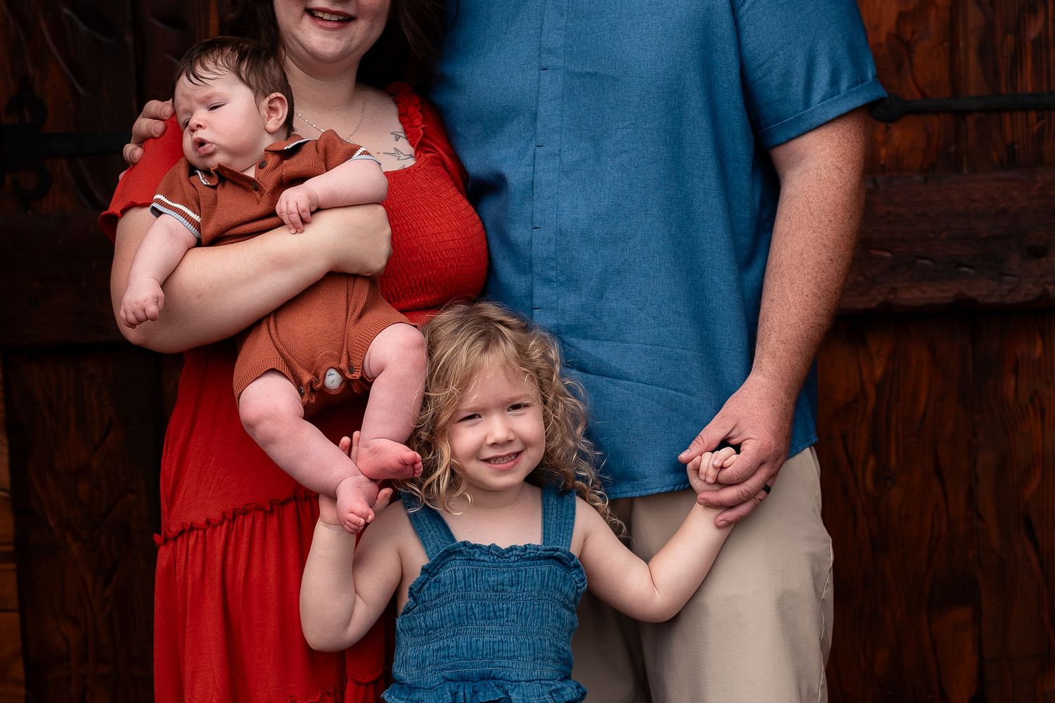 Family portrait with mother in red dress holding infant, father in blue shirt, and toddler girl in denim overalls smiling.