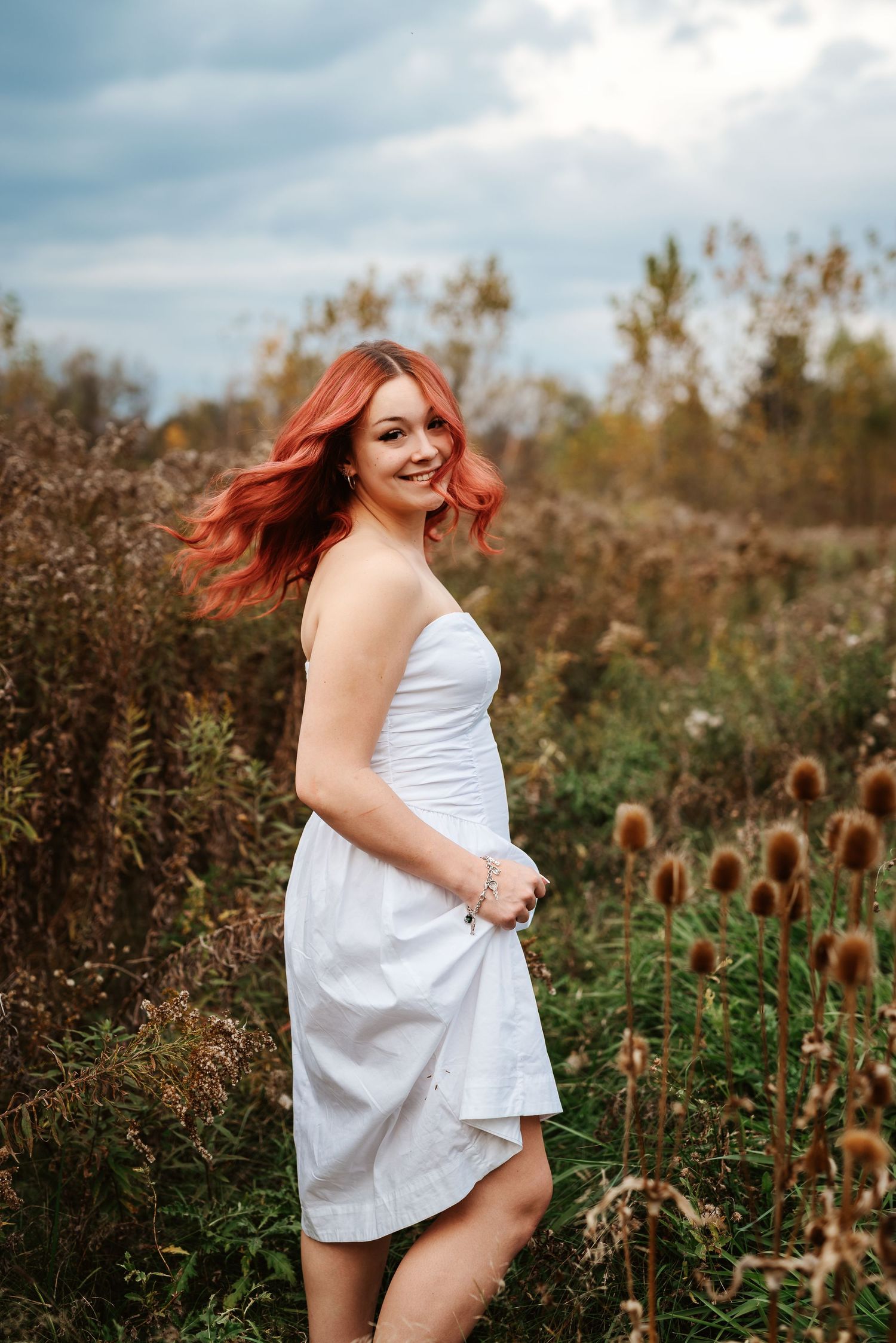Young woman with red hair in white dress posing in autumn field with wildflowers under cloudy sky.