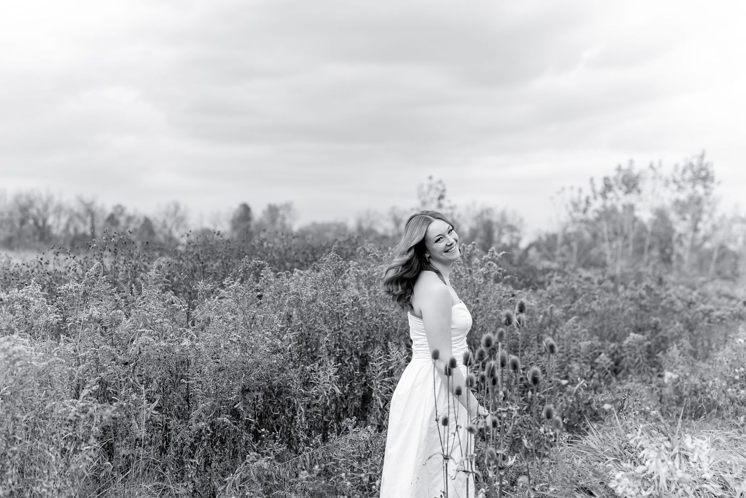 Black and white photo of woman in white dress standing among wildflowers in an open field.