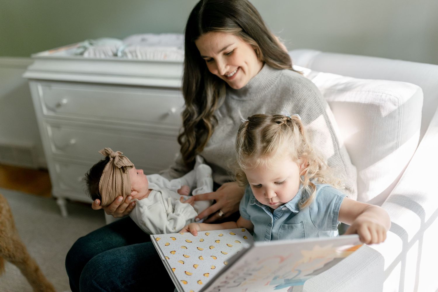 Mother reads picture book with toddler girl and newborn baby in nursery chair.