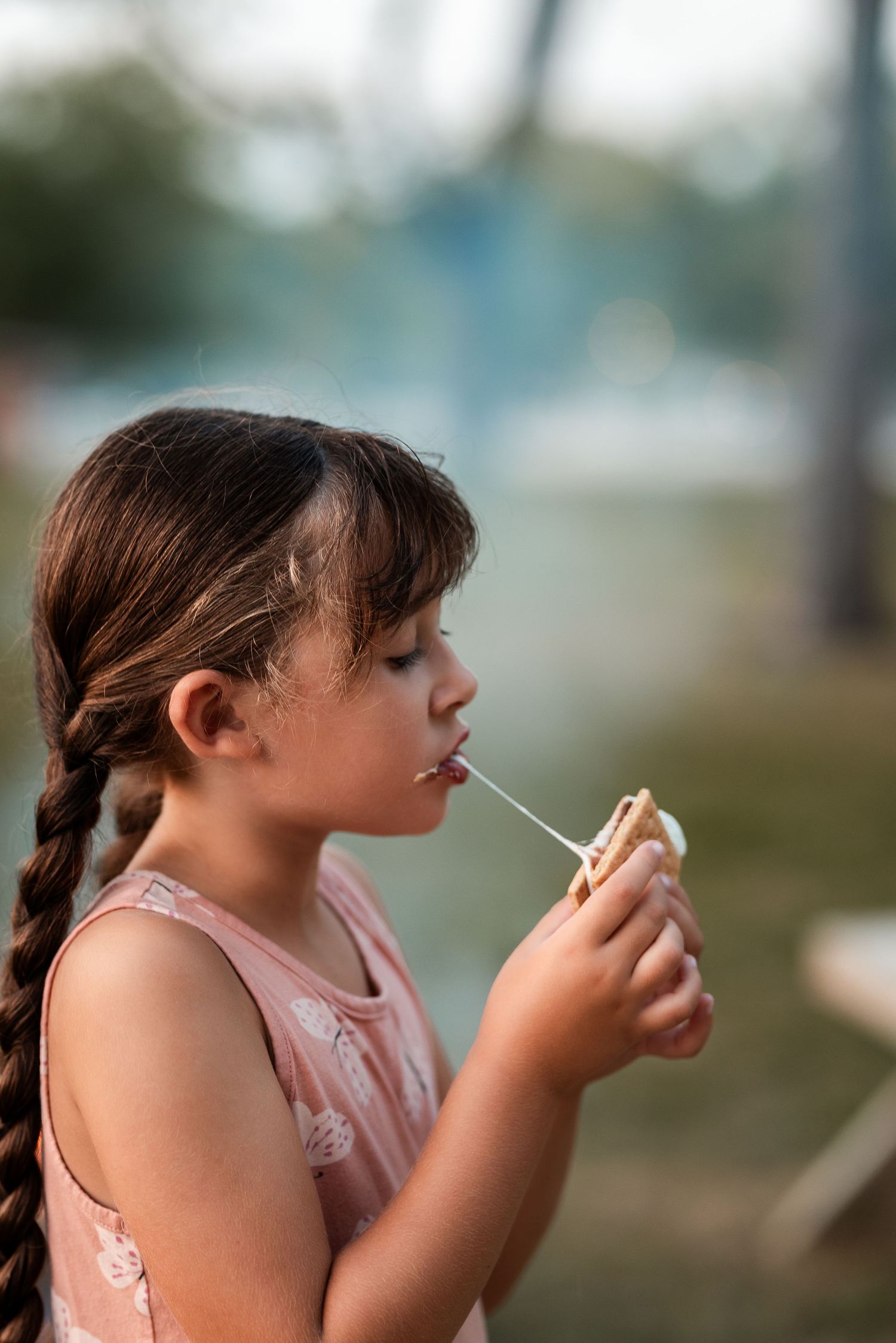 Young girl with braided hair eating a s'more on a stick outdoors, wearing a pink sleeveless top.