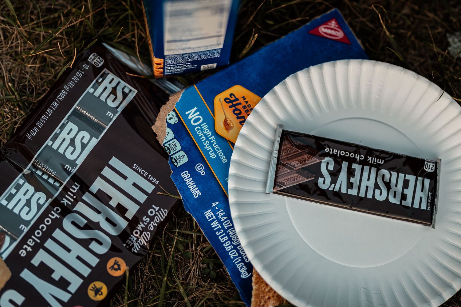 Hersheys milk chocolate bars and wrappers scattered on grass next to a white paper plate outdoors.