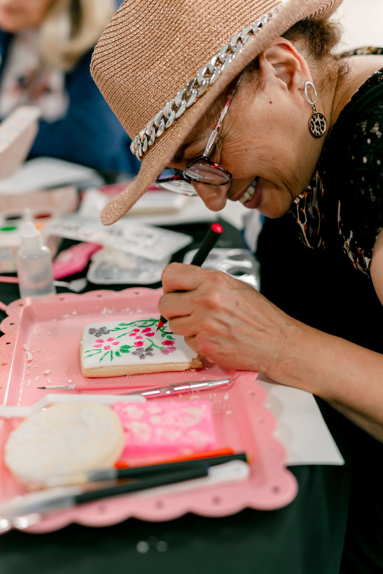 Elderly woman in straw hat decorating a floral cookie with icing at a cookie decorating class.
