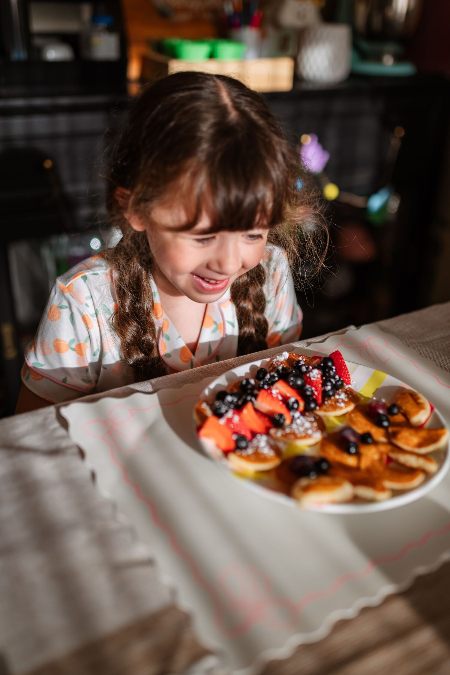 Young child with braids smiling at a plate of colorful decorated donuts on a white tablecloth.