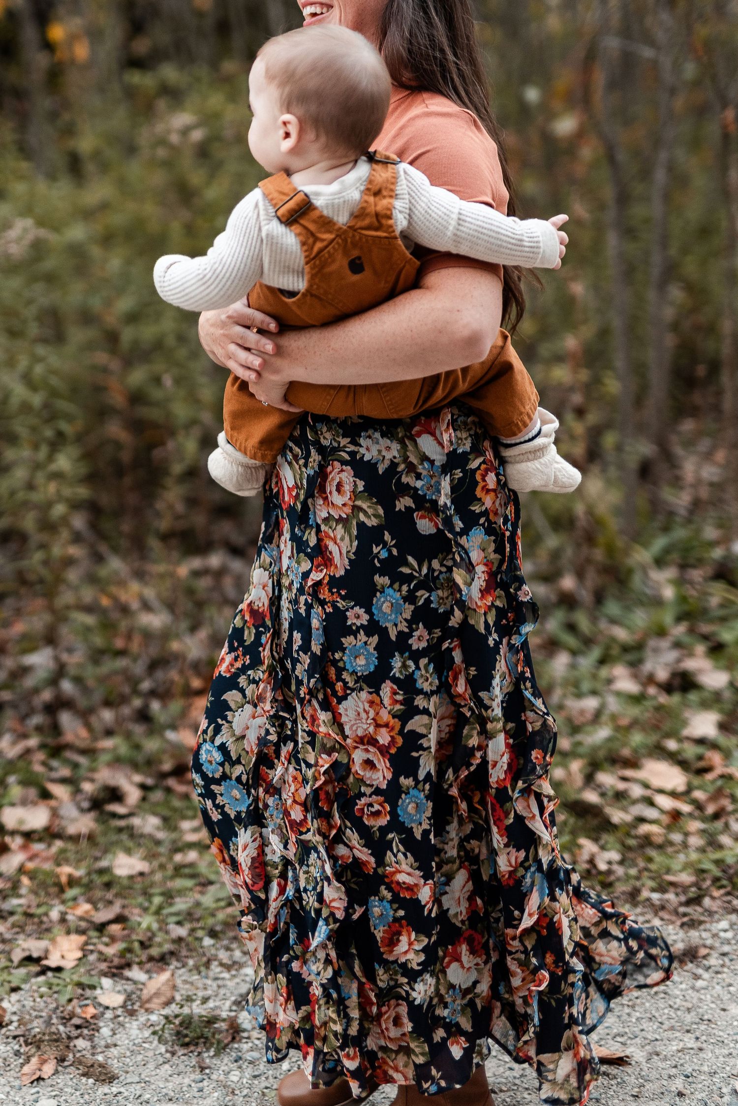 Woman in a floral maxi skirt carries a baby in rust corduroy overalls along a leaf-covered woodland path in autumn.