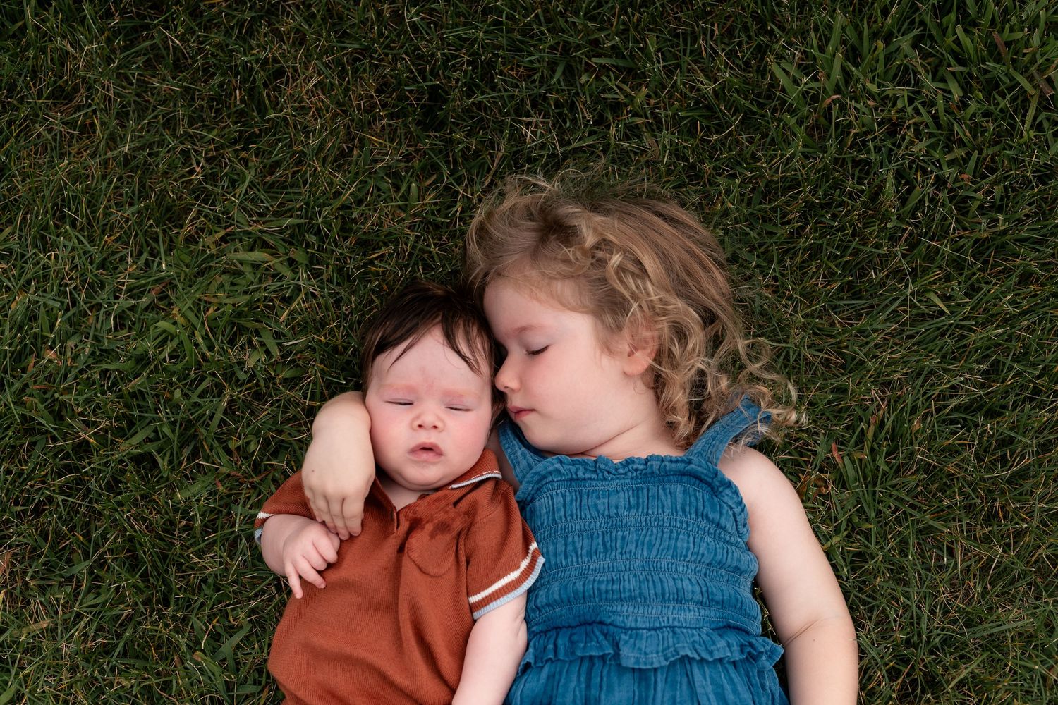 Toddler girl in blue dress cuddling sleeping newborn baby on green grass outdoors.