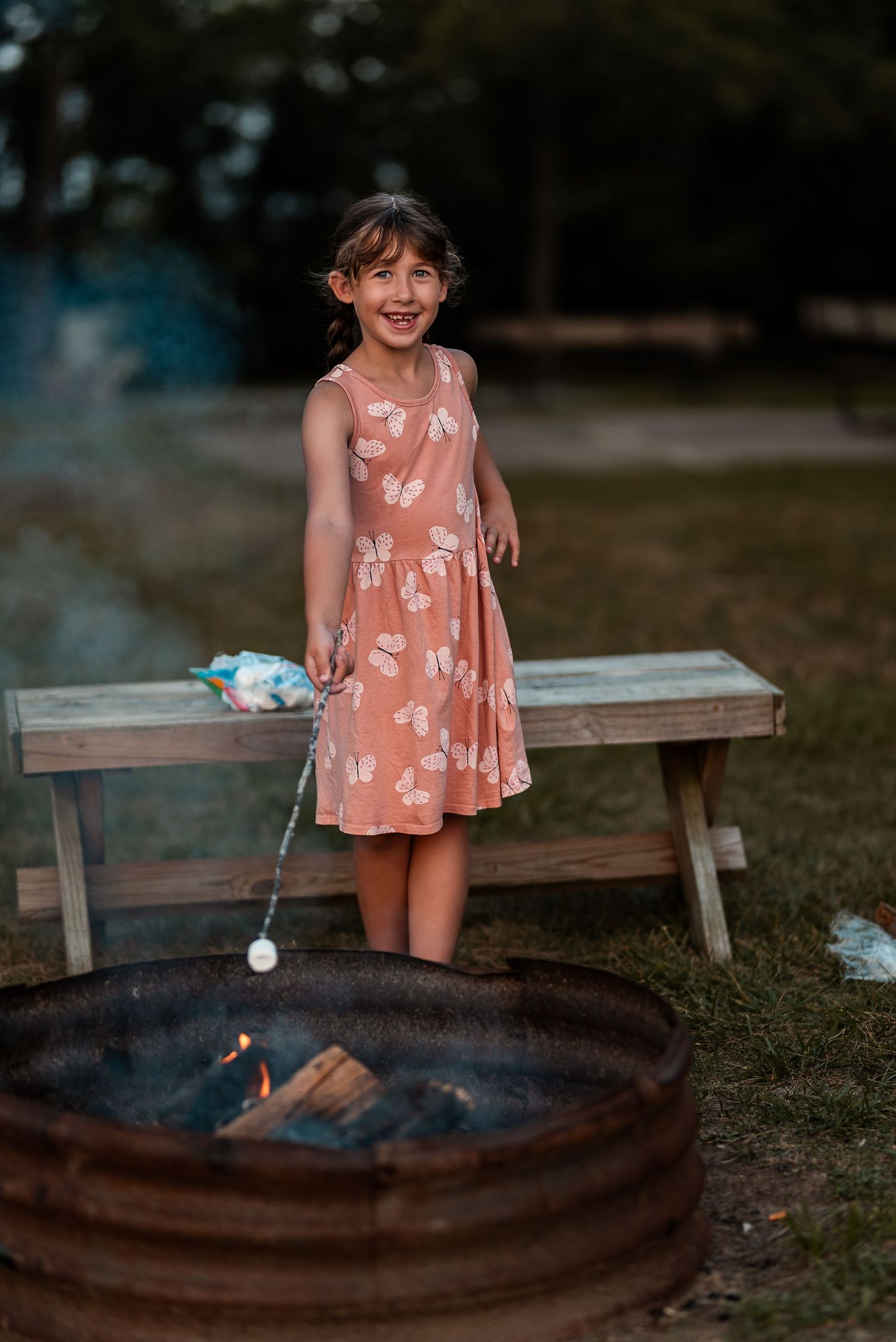 A smiling young girl in a pink floral dress roasting a marshmallow over a campfire outdoors.