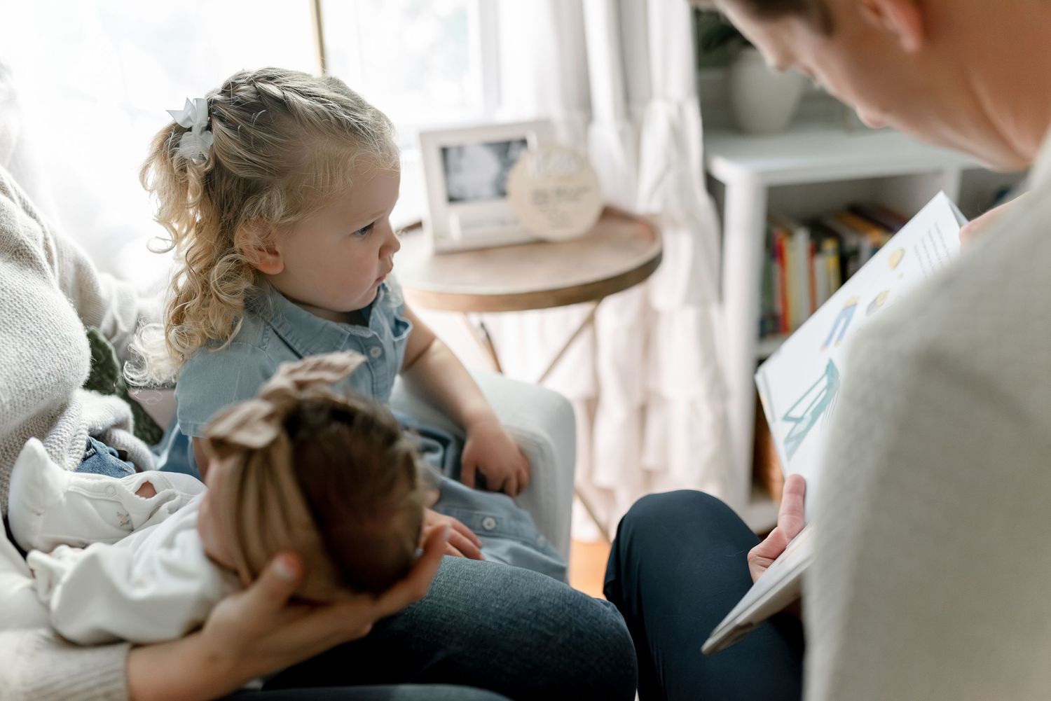 A mother reads a book to her toddler daughter while holding a newborn baby on a cozy chair.