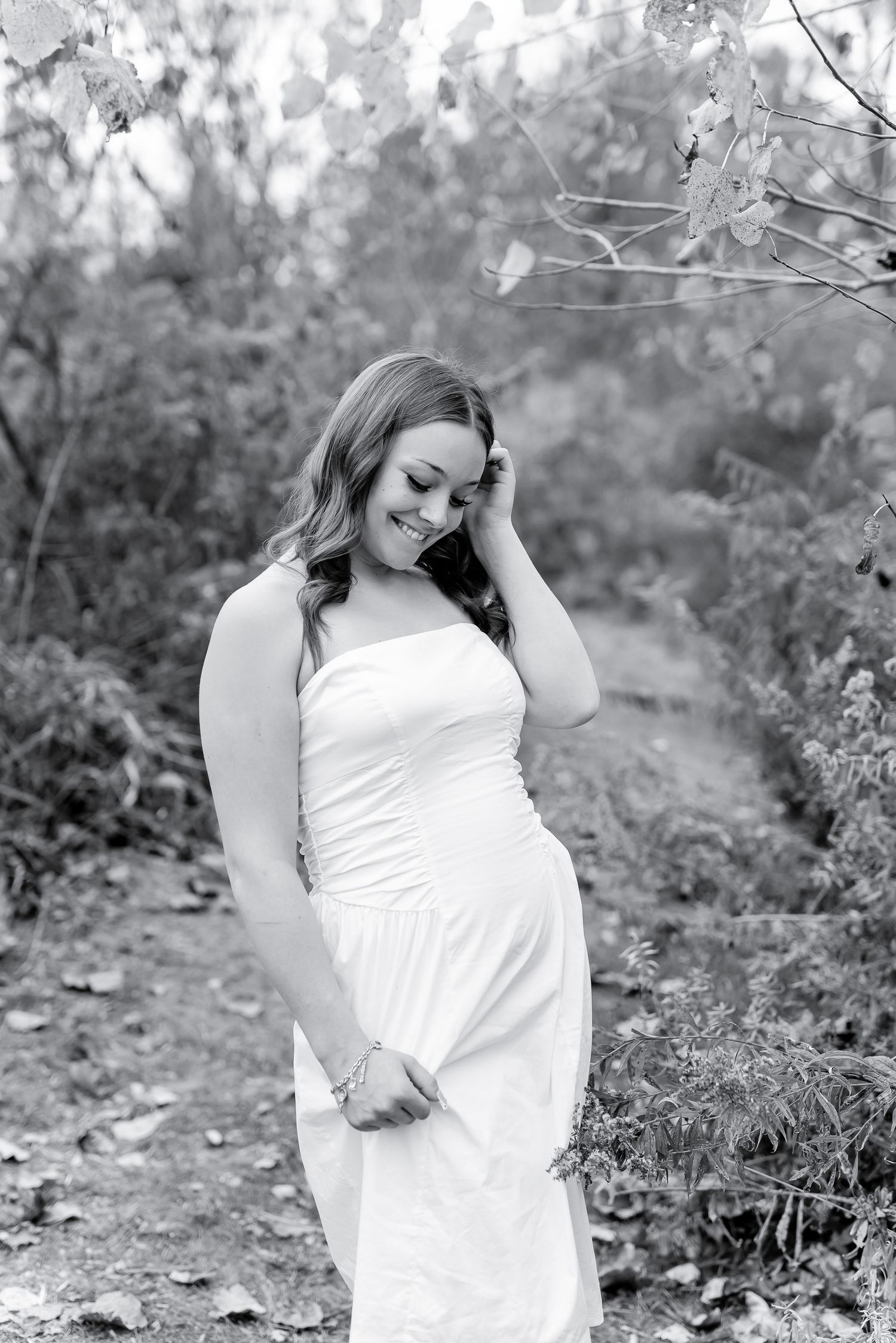 Woman in white strapless dress smiling shyly in black and white outdoor portrait.