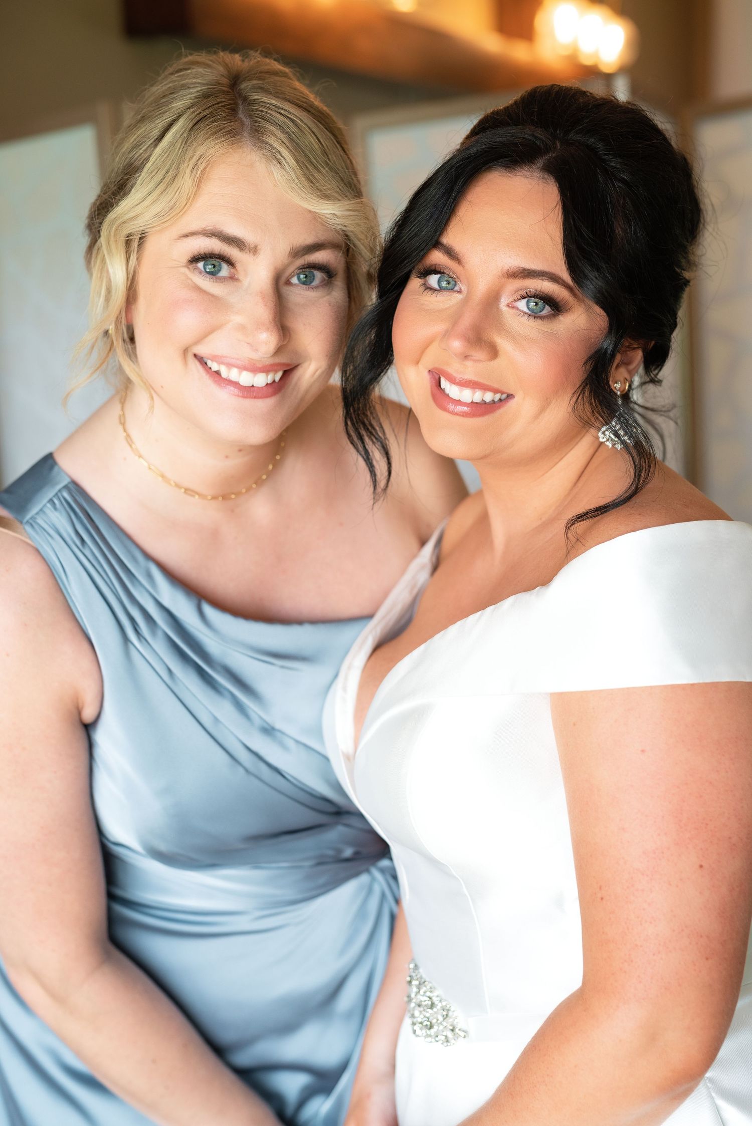 Two women pose closely together, with one wearing a light blue bridesmaid dress and the other in a white bridal gown.
