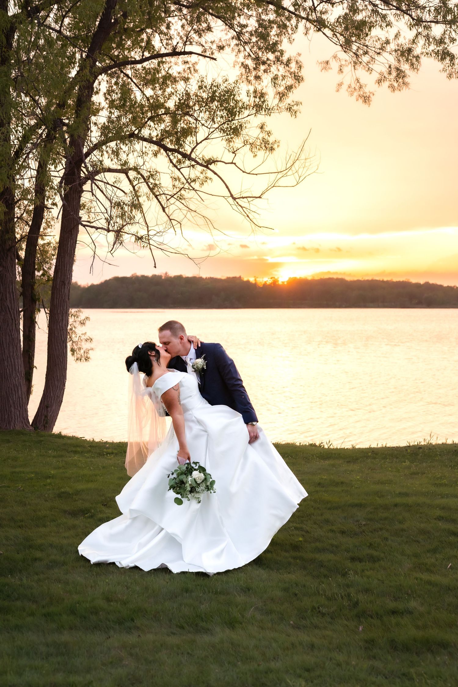 Elegant wedding couple shares romantic dip pose at sunset by serene lake with tree silhouette in foreground.