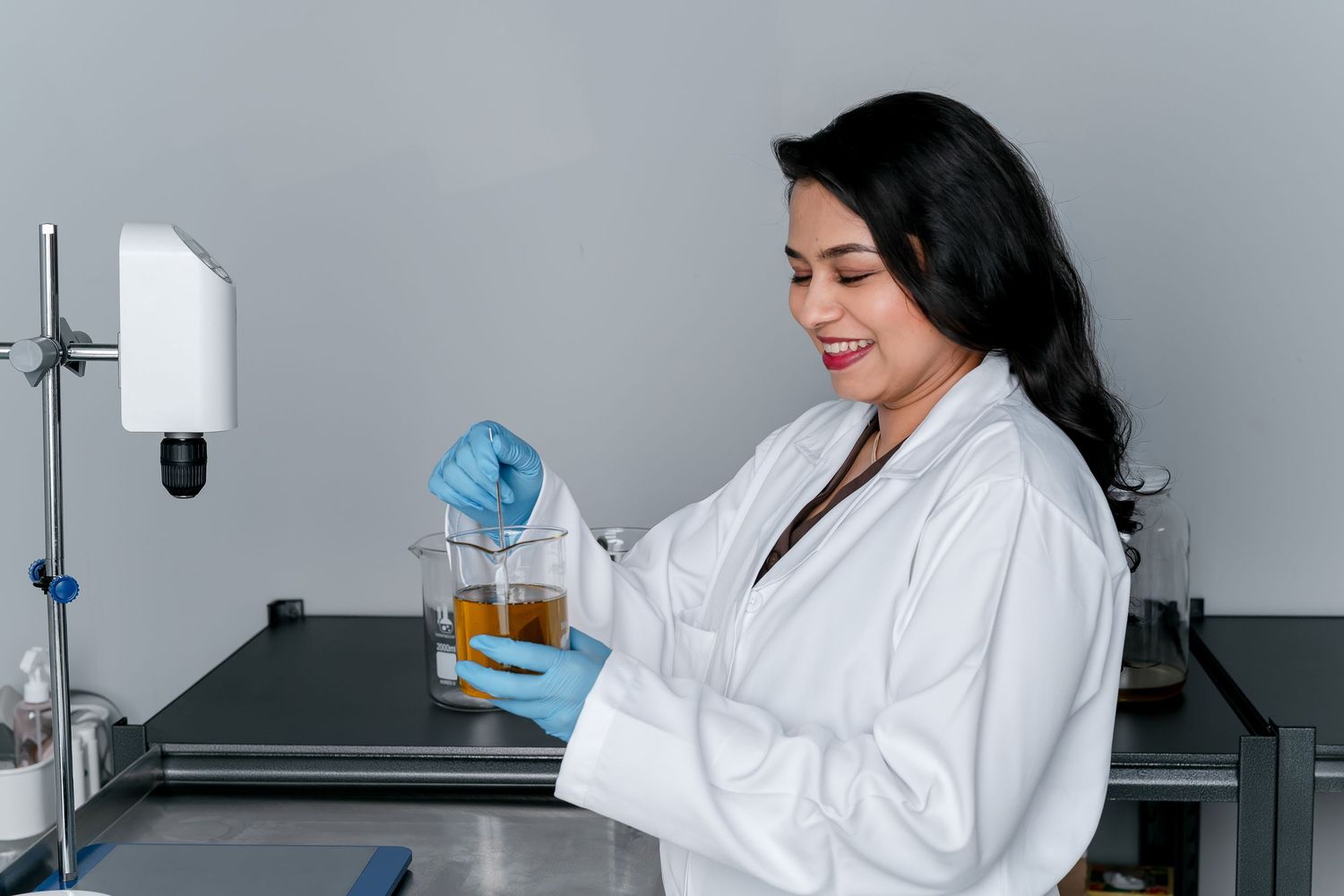 Female scientist in white lab coat and blue gloves smiling while examining amber liquid in beaker in laboratory.